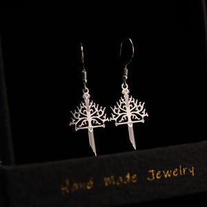 May include: A pair of silver-colored earrings in a black jewelry box. Each earring features a tree-like design atop a sword-shaped pendant. The box is labeled "Hand Made Jewelry" in gold lettering.