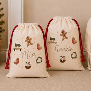 May include: Two off-white canvas drawstring bags with red cords, featuring printed illustrations of gingerbread people, cars with trees, robins, wreaths, and Christmas puddings. The bags are personalised with the names Mia and Frankie in script.