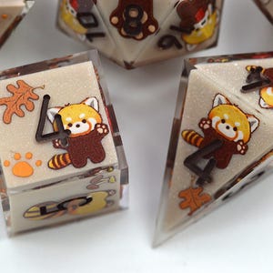 May include: A set of multi-sided resin dice featuring a beige glitter base with embedded illustrations of red pandas, orange oak leaves, and paw prints. Each die face displays a dark brown numeral, with the red panda characters integrated into the design.