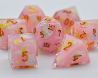 Op de afbeelding: Set van zeven roze dobbelstenen met gouden cijfers en witte illustraties van een konijn, maan en sterren. De dobbelstenen zijn gemaakt van een doorschijnend materiaal met een glinsterende afwerking.