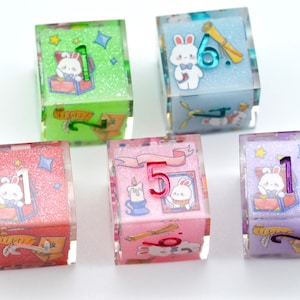 May include: Six colorful dice with a glittery finish. Each die features a different number from 1 to 6 and a cartoon bunny rabbit illustration. The dice are pink, green, blue, purple, and white.