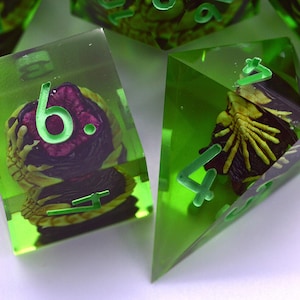 Puede incluir: Dados poliédricos de resina verde lima translúcida con motivos de manos esqueléticas incrustados en color morado oscuro y amarillo. Los dados tienen bordes afilados y números grabados en verde brillante. Se ven un dado de cuatro y seis caras.