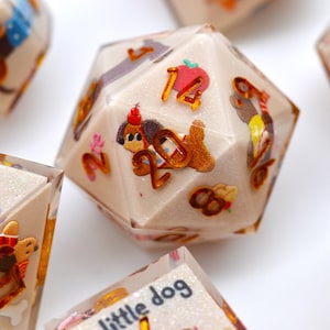 Puede incluir: Primer plano de varios dados poliédricos con base color crema. Cada dado presenta ilustraciones coloridas de perros de dibujos animados, números y otros diseños lúdicos. Los dados están esparcidos sobre una superficie blanca.