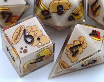 Puede incluir: Varios dados poliédricos con exterior de resina transparente e interior beige. Cada dado presenta un número e ilustraciones de carpinchos, alimentos y la palabra "capybare". Los dados están diseñados para juegos de mesa.