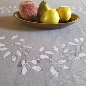 May include: A gray tablecloth featuring a white printed leaf vine pattern. An oval-shaped brown bowl sits on the fabric, containing one textured red apple and three smooth yellow pears arranged as a decorative centerpiece on the table.