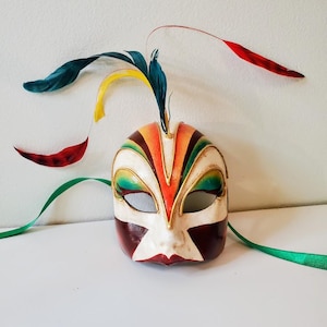 May include: A decorative Venetian mask with a white base, adorned with colorful stripes in orange, brown, and green. Gold trim outlines the eye openings. Feathers in red, yellow, and teal extend from the top, with green ribbons.
