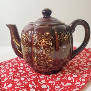 May include: A brown ceramic teapot with a lid and handle, decorated with gold floral patterns and colorful accents. The teapot sits on a red floral patterned cloth. The spout and rim have gold trim.