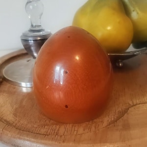 May include: A shiny, reddish-orange egg-shaped object with small holes, possibly a salt or pepper shaker, sits on a wooden tray. In the background are two yellow fruits and a silver-based glass object.
