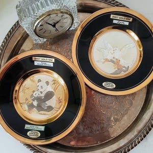 May include: Two decorative plates with 24kt gold rims and black borders. One plate features a panda and cub with the text Earth Day Wildlife Series Collection. The other plate shows two white doves with the text Love. A crystal clock sits nearby.