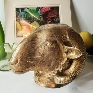 May include: A metallic gold-toned ram head sculpture featuring detailed, textured fur patterns and large, curved horns. The sculpture is cast in a solid, reflective material with a smooth finish on the snout and ears, capturing the animal's profile.