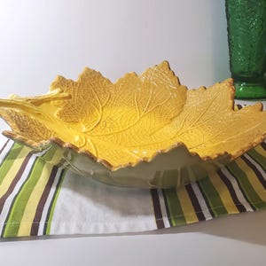 May include: A large, yellow ceramic leaf-shaped bowl with detailed veining. The bowl sits on a striped green, brown, and white cloth. The bowl is a decorative piece, perfect for serving or display.