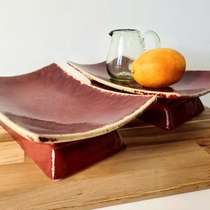 May include: Two rectangular ceramic dishes with a deep maroon glaze and cream-colored edges. The dishes feature a curved, elevated design. A small, clear glass pitcher and a single yellow lemon are placed on the rear dish, showcasing the set.