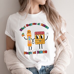 May include: White t-shirt with a colorful graphic of a bingo card and a bingo dauber. The text "What Happens at Bingo Stays at Bingo" is printed on the shirt.