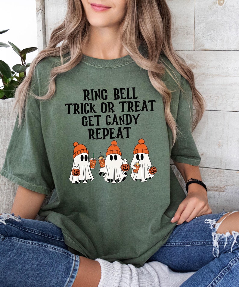 Może przedstawiać: Zielona koszulka w kolorze oliwkowym z napisem "RING BELL TRICK OR TREAT GET CANDY REPEAT". Koszulka przedstawia grafikę trzech duch&oacute;w w pomarańczowych czapkach trzymających dynie i napoje. Koszulka jest w wygodnym, swobodnym stylu.