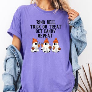 Może przedstawiać: Fioletowa koszulka z napisem "RING BELL TRICK OR TREAT GET CANDY REPEAT". Koszulka przedstawia trzy kresk&oacute;wkowe duszki w pomarańczowych czapkach, trzymające dynie i napoje. Swobodny design z motywem Halloween.