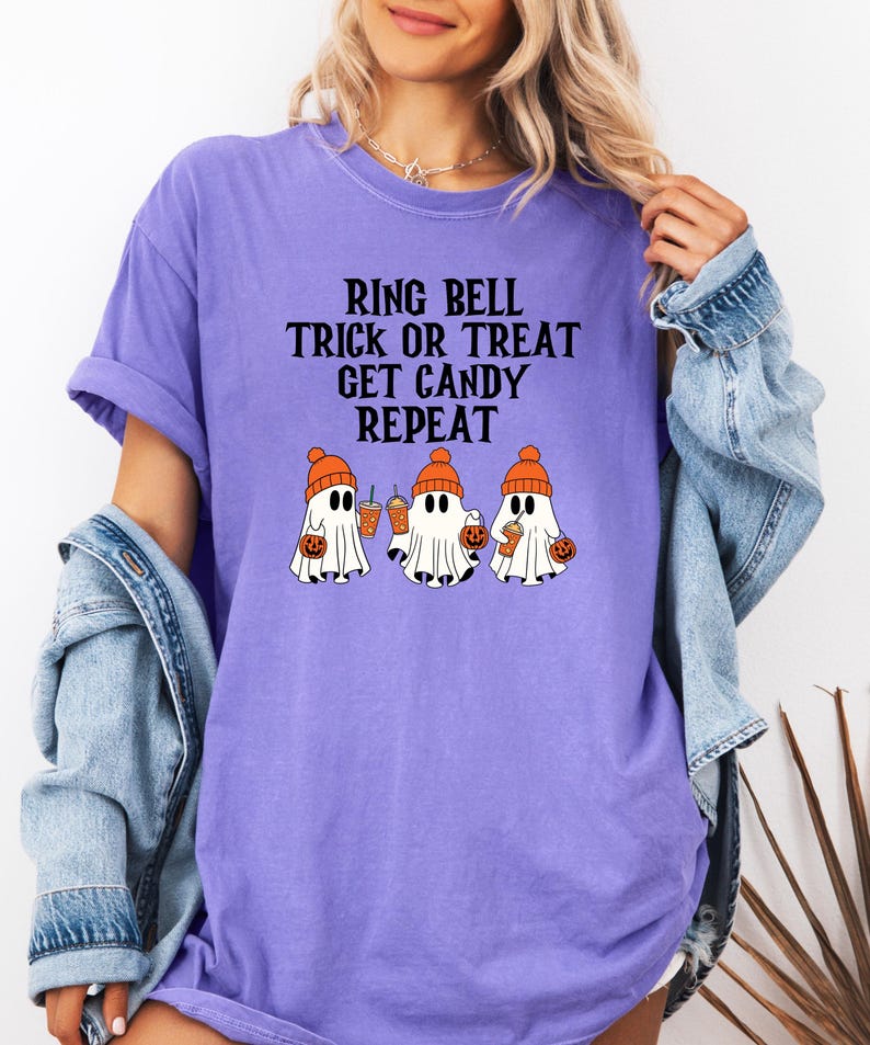 Może przedstawiać: Fioletowa koszulka z napisem "RING BELL TRICK OR TREAT GET CANDY REPEAT". Koszulka przedstawia trzy kresk&oacute;wkowe duszki w pomarańczowych czapkach, trzymające dynie i napoje. Swobodny design z motywem Halloween.