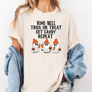Może przedstawiać: Kremowy t-shirt z czarnym napisem "RING BELL TRICK OR TREAT GET CANDY REPEAT". Poniżej znajdują się trzy kresk&oacute;wkowe duszki w pomarańczowych czapkach, trzymające dynie i napoje. Koszulka ma kr&oacute;tkie rękawy i okrągły dekolt.