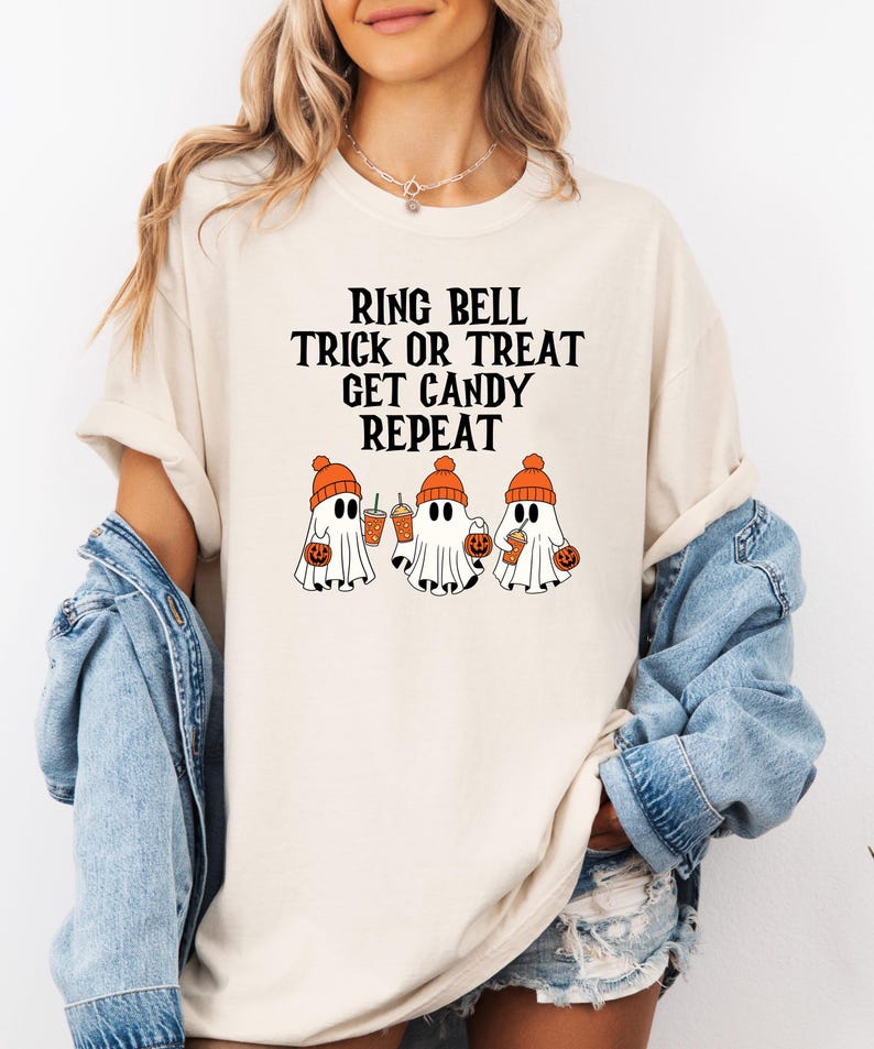 Może przedstawiać: Kremowy t-shirt z czarnym napisem "RING BELL TRICK OR TREAT GET CANDY REPEAT". Poniżej znajdują się trzy kresk&oacute;wkowe duszki w pomarańczowych czapkach, trzymające dynie i napoje. Koszulka ma kr&oacute;tkie rękawy i okrągły dekolt.
