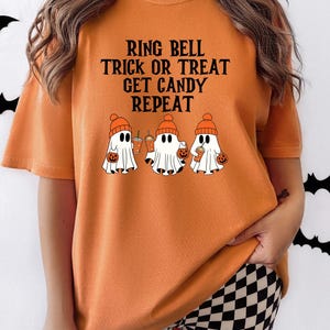 Może przedstawiać: Pomarańczowa koszulka z napisem "RING BELL TRICK OR TREAT GET CANDY REPEAT". Koszulka przedstawia trzy kresk&oacute;wkowe duszki w pomarańczowych czapkach, trzymające torby na słodycze w kształcie dyni i napoje. Motyw Halloween.