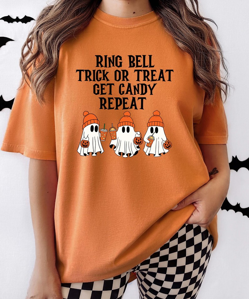 Może przedstawiać: Pomarańczowa koszulka z napisem "RING BELL TRICK OR TREAT GET CANDY REPEAT". Koszulka przedstawia trzy kresk&oacute;wkowe duszki w pomarańczowych czapkach, trzymające torby na słodycze w kształcie dyni i napoje. Motyw Halloween.