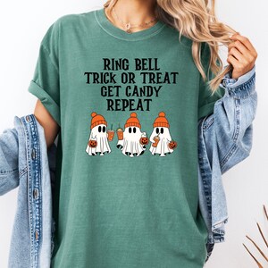 Może przedstawiać: Zielona koszulka z napisem "RING BELL TRICK OR TREAT GET CANDY REPEAT". Koszulka przedstawia grafikę trzech kresk&oacute;wkowych duch&oacute;w w pomarańczowych czapkach, trzymających dynie i napoje, idealna na Halloween.