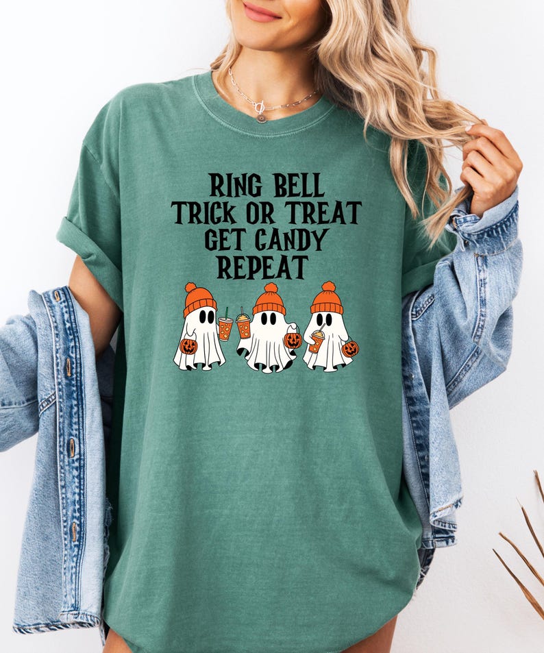 Może przedstawiać: Zielona koszulka z napisem "RING BELL TRICK OR TREAT GET CANDY REPEAT". Koszulka przedstawia grafikę trzech kresk&oacute;wkowych duch&oacute;w w pomarańczowych czapkach, trzymających dynie i napoje, idealna na Halloween.