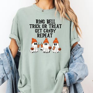 Może przedstawiać: Jasnozielona koszulka z napisem "RING BELL TRICK OR TREAT GET CANDY REPEAT". Koszulka przedstawia grafikę trzech duch&oacute;w w pomarańczowych czapkach, trzymających dynie i napoje. Koszulka jest w wygodnym, swobodnym stylu.