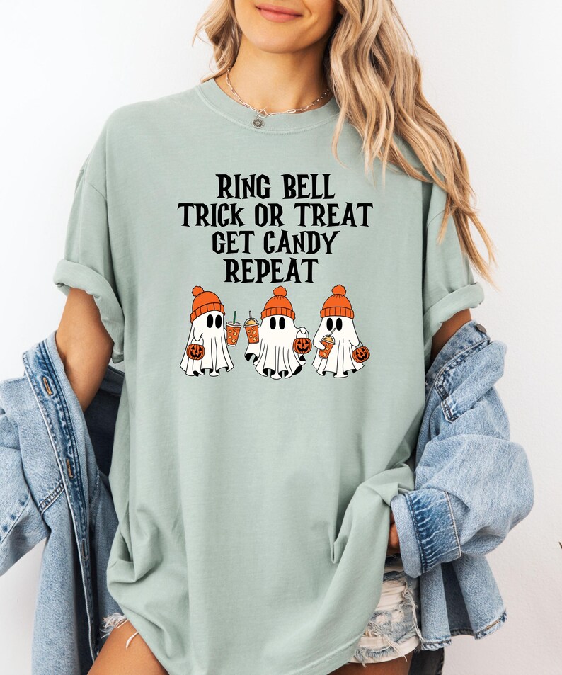 Może przedstawiać: Jasnozielona koszulka z napisem "RING BELL TRICK OR TREAT GET CANDY REPEAT". Koszulka przedstawia grafikę trzech duch&oacute;w w pomarańczowych czapkach, trzymających dynie i napoje. Koszulka jest w wygodnym, swobodnym stylu.