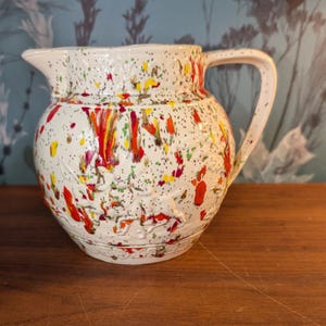 Peut inclure: Une cruche en céramique de couleur crème avec un motif en relief représentant des cerfs et du feuillage. La surface est décorée d'un motif d'éclaboussures de peinture aux tons rouge, jaune et vert, avec une anse courbée et un bec verseur.