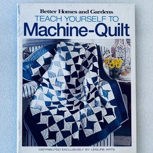 May include: A softcover instructional book titled Better Homes and Gardens Teach Yourself to Machine-Quilt. The cover features a blue and white geometric quilt draped over a striped chair, with a vase of yellow flowers and a telephone nearby.