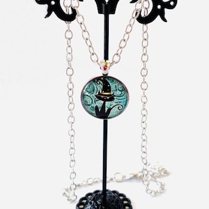 May include: A silver-toned chain necklace featuring a round pendant with a glass cabochon. The pendant displays a black cat illustration wearing a pointed hat against a teal, swirling background. The necklace is displayed on a black metal stand.