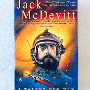 May include: A paperback book titled A Talent for War by Jack McDevitt. The cover features a bearded person in a space helmet against a vibrant orange and blue nebula background, with a small spaceship flying below and text by Stephen King.