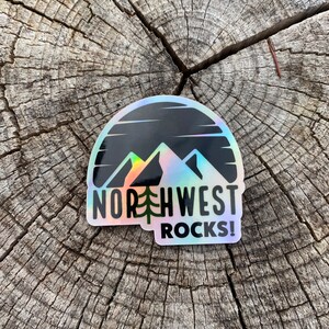 May include: A holographic sticker with a black and white mountain silhouette and a sunset. The text "NORTHWEST ROCKS!" is written in black below the mountains.