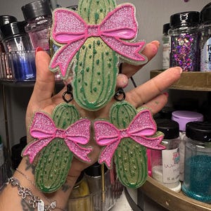 May include: Green pickle-shaped earrings with pink glitter bows. The earrings have a textured surface and a loop at the top for hanging. The background includes jars of glitter and other craft supplies.