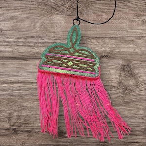 May include: A green and pink layered tassel ornament with a black cord. The ornament has a geometric design and is decorated with glitter. The tassel is made of pink yarn.