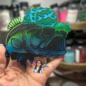 May include: A colorful, stylized fish-shaped item with a cap. The fish is primarily black, green, and blue, with a gradient effect. The cap is green and blue. The item appears to be a decorative piece or a craft item.