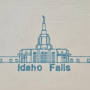 May include: Embroidered design of the Idaho Falls Temple, a white building with a tall spire, outlined in blue thread on a white fabric background. The text "Idaho Falls" is embroidered below the building.