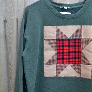 May include: A dark green sweatshirt with a patchwork quilt design featuring a red and black plaid star in the center. The quilt design is sewn onto the front of the sweatshirt.