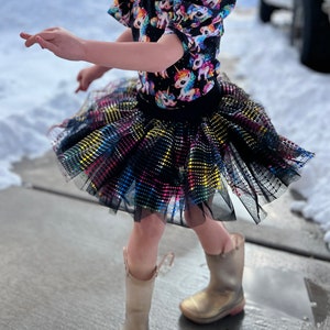 May include: A child's outfit featuring a black top with a unicorn pattern and a multi-colored dot tulle skirt. The outfit is paired with gold boots. The child is outside on a snowy day.