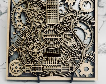 May include: Wooden wall art featuring a detailed laser-cut design of a guitar integrated with gears and mechanical elements. The artwork is set against a silver background within a square wooden frame. The piece is displayed on a black stand.