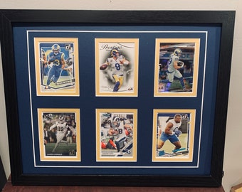 Könnte beinhalten: Eine gerahmte Präsentation von sechs Football-Sammelkarten mit Spielern der Los Angeles Rams. Die Karten befinden sich in einem schwarzen Rahmen mit einer blauen Passepartout und goldenen Akzenten. Die gezeigten Spieler sind Aaron Donald, Cooper Kupp, Tyler Higbee, Matthew Stafford und zwei weitere Spieler.