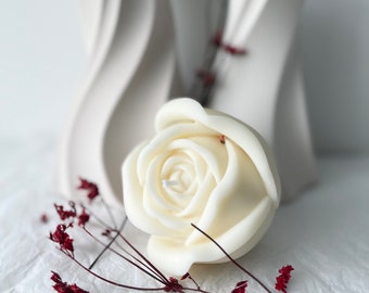 Può includere: Una candela bianca a forma di rosa con un design delicato e realistico. La candela è appoggiata su una superficie bianca con fiori rossi secchi.