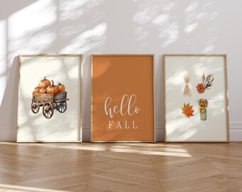 May include: Three framed wall art prints. The left print shows a wooden wagon filled with orange pumpkins. The center print has a solid orange background with the white text "hello FALL". The right print features botanical illustrations.