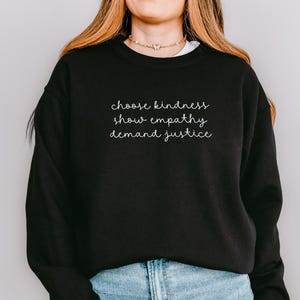 May include: A black crewneck sweatshirt featuring white cursive text on the chest that reads choose kindness, show empathy, demand justice. The garment has long sleeves and a ribbed neckline, paired with light blue denim jeans.