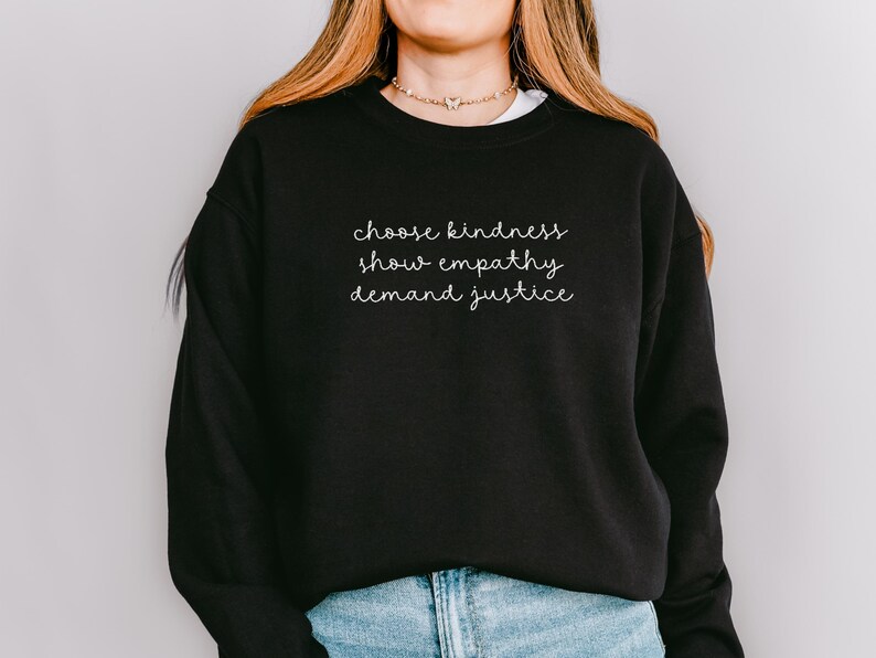 May include: A black crewneck sweatshirt featuring white cursive text on the chest that reads choose kindness, show empathy, demand justice. The garment has long sleeves and a ribbed neckline, paired with light blue denim jeans.