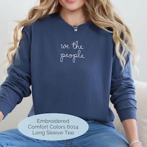 May include: A navy blue long-sleeve crewneck t-shirt featuring the white embroidered text "we the people" centered on the chest. The garment is a Comfort Colors 6014 model, characterized by its relaxed fit and long sleeves.