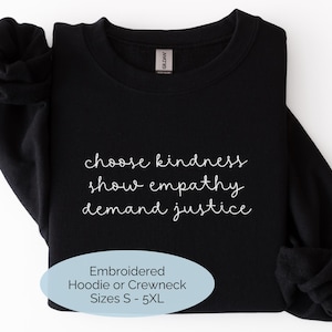 May include: A black long-sleeve crewneck sweatshirt featuring white embroidered text that reads choose kindness, show empathy, demand justice. A light blue oval graphic overlay at the bottom includes the text Embroidered Hoodie or Crewneck Sizes S - 5XL.