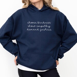 May include: A navy blue hooded sweatshirt featuring a front kangaroo pocket and white cursive text printed on the chest that reads: choose kindness, show empathy, demand justice. The garment has long sleeves and a hood with matching drawstrings.