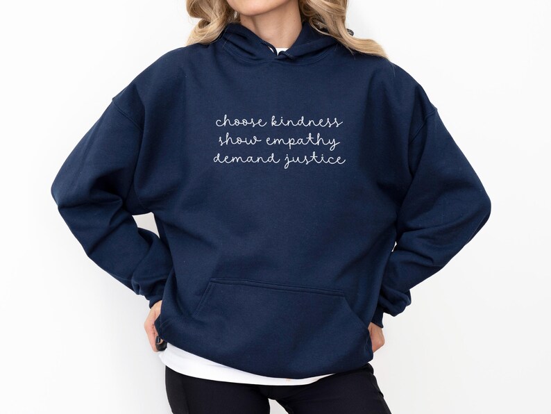 May include: A navy blue hooded sweatshirt featuring a front kangaroo pocket and white cursive text printed on the chest that reads: choose kindness, show empathy, demand justice. The garment has long sleeves and a hood with matching drawstrings.