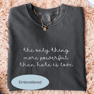May include: A dark gray t-shirt with the embroidered text "the only thing more powerful than hate is love." A gold chain necklace with a heart pendant rests on the shirt. The shirt is folded on a fluffy, light pink surface.